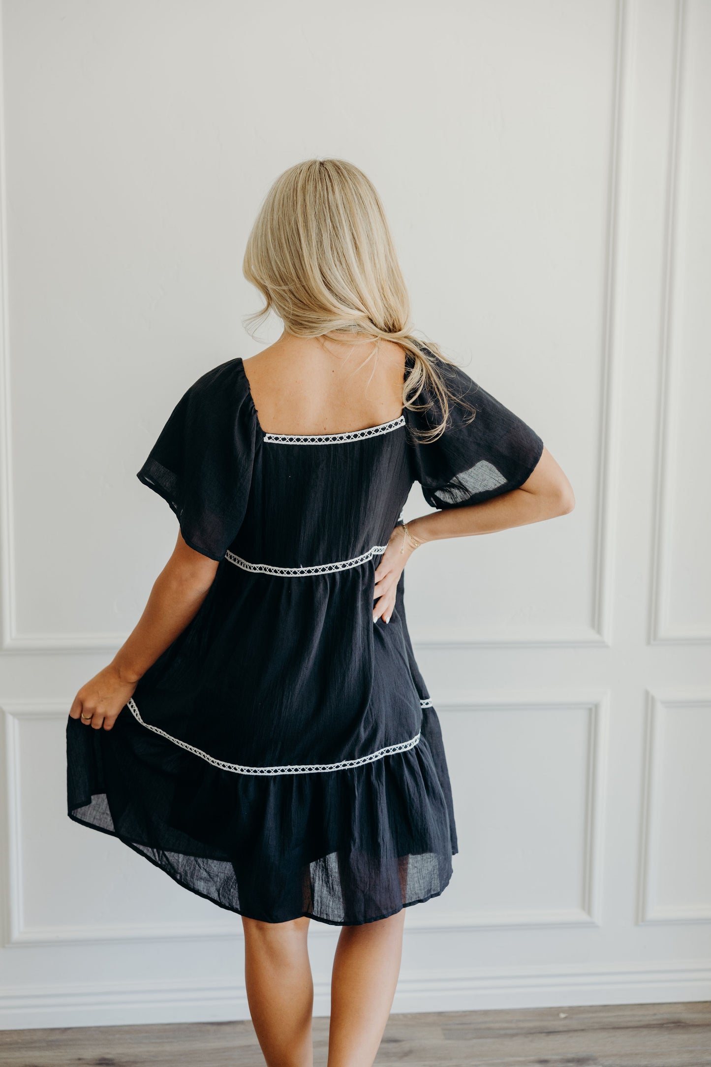 Back of knee length black dress with with tiers, white lace trim, square neckline.