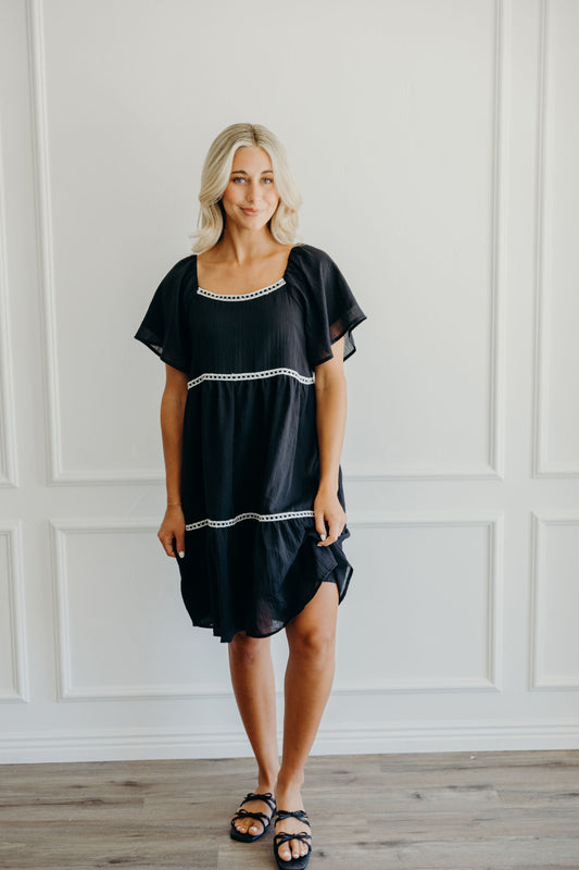 Knee length black tiered dress with white lace detail, square neckline, and flutter sleeves.